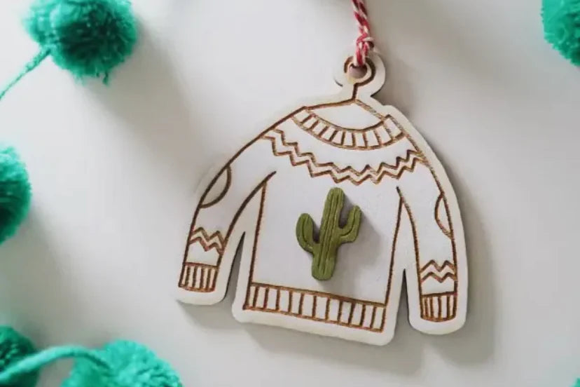 Decorative ornament shaped like a sweater with a cactus on a string of green pom-poms.